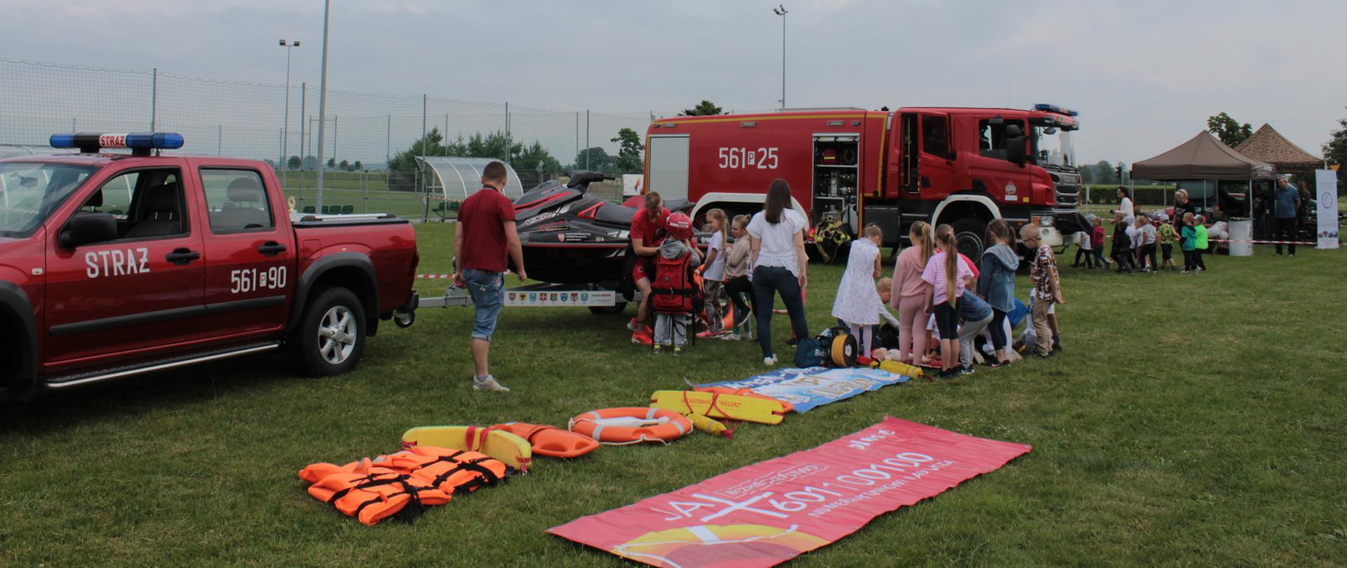 Na pierwszym planie dzieciu uczą się podstaw udzielania pierwszej pomocy, dalej stoi wóz strażacki, który ogląda kolejn grupa uczniów, ze nimi idoczne stanowiska edukacyjne Wojska Polskiego