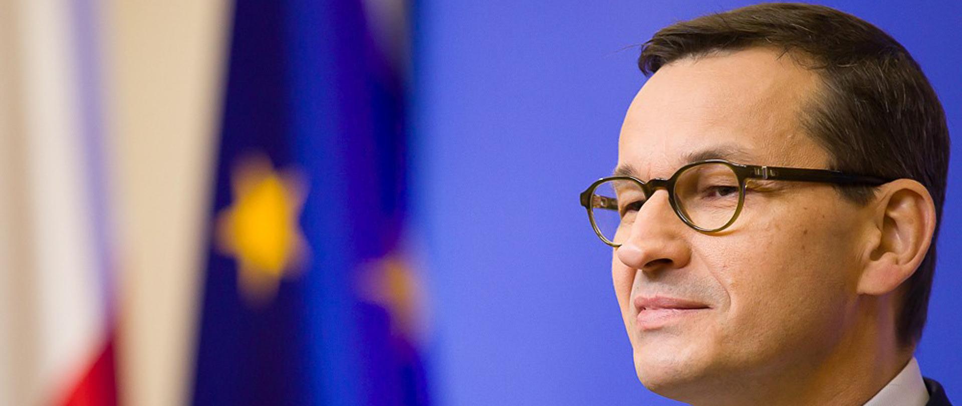 Premier Mateusz Morawiecki, w tle flaga Polski i flaga UE. 