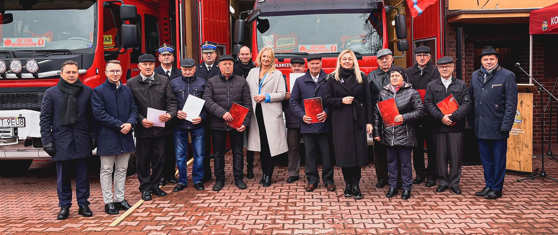 Parlamentarzyści, funkcjonariusze Państwowej Straży Pożarnej oraz druhowie, którzy otrzymali świadczenia ratownicze stoją przed budynkiem remizy Ochotniczej Straży Pożarnej w Ostrzeszowie.