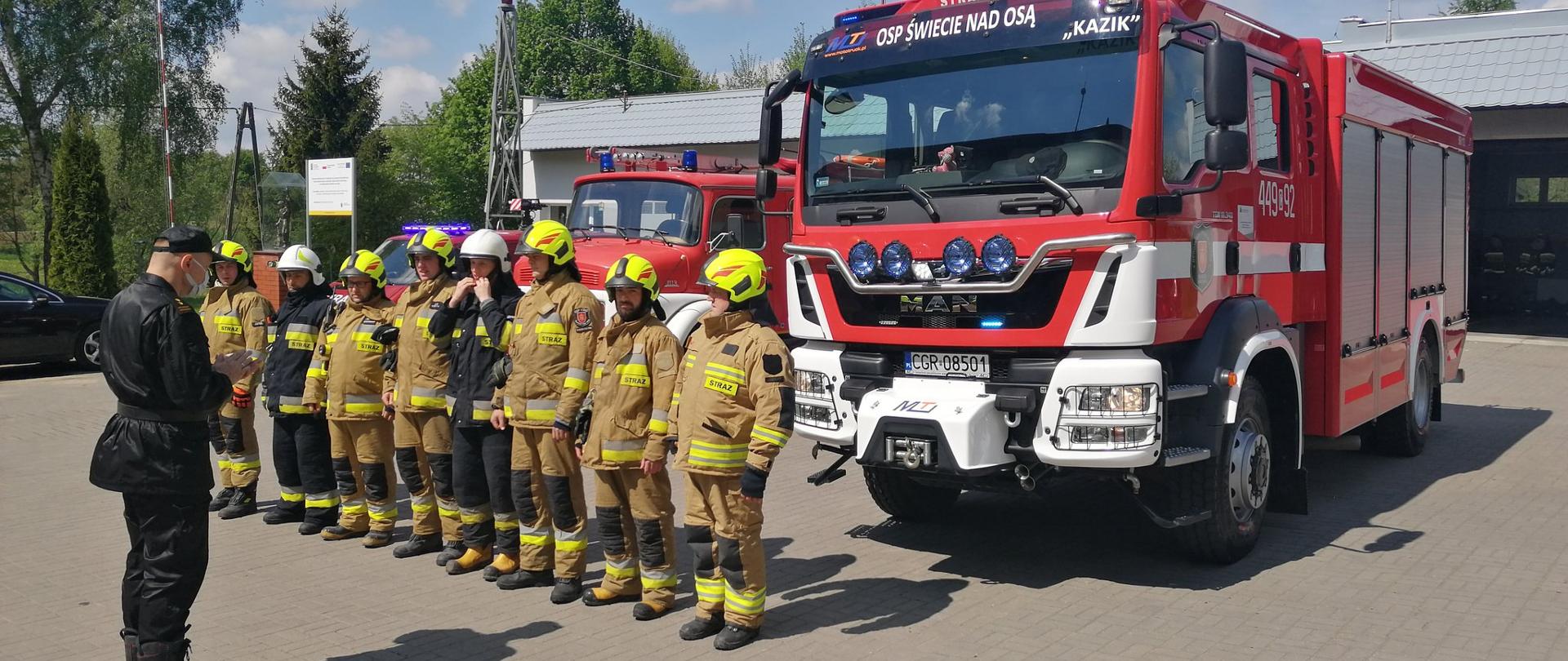 Zdjęcie przedstawia 3 czerwono-biało-srebrne wozy strażackie. Przed nimi w szeregu stoją strażacy w piaskowych i czarnych mundurach bojowych. Naprzeciw nich strażak w czarnym mundurze sztabowym przeprowadza inspekcję.