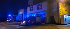 Dwa samochody pożarnicze wysunięte przed bramy garażowe. Maja włączone światła błyskowe. Za nimi budynek remizy. Sceneria nocna. 