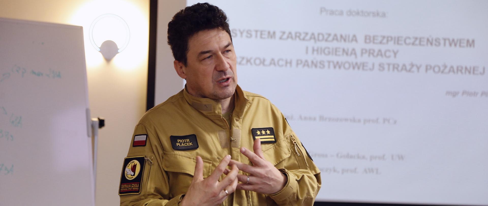 Komendant Centralnej Szkoły PSP podczas wystąpienia, w tle ekran multimedialny z wyświetlonym slajdem prezentacji