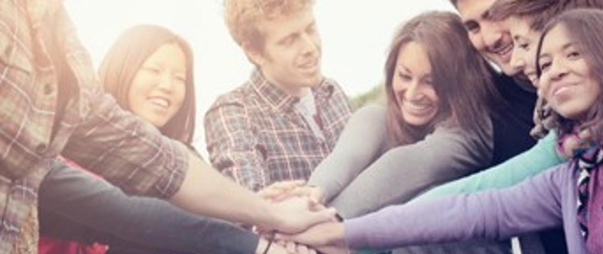 Grupa 7 młodych, uśmiechniętych ludzi - mężczyzn i kobiet, stojąca w kręgu z jedną ręką skierowaną do środka. Dłonie wszystkich połączone - ułożone jedna na drugiej.