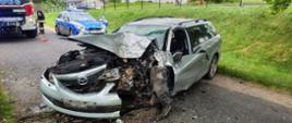 Na wiejskiej drodze stoi poważnie uszkodzony samochód osobowy typu kombi po wypadku drogowym. Przód pojazdu jest całkowicie zniszczony, szczególnie po stronie kierowcy, z widocznymi uszkodzeniami silnika i karoserii. Na jezdni leżą fragmenty rozbitych elementów auta. W tle znajdują się samochód straży pożarnej, radiowóz policyjny oraz zabudowania mieszkalne. Niebo jest zachmurzone.