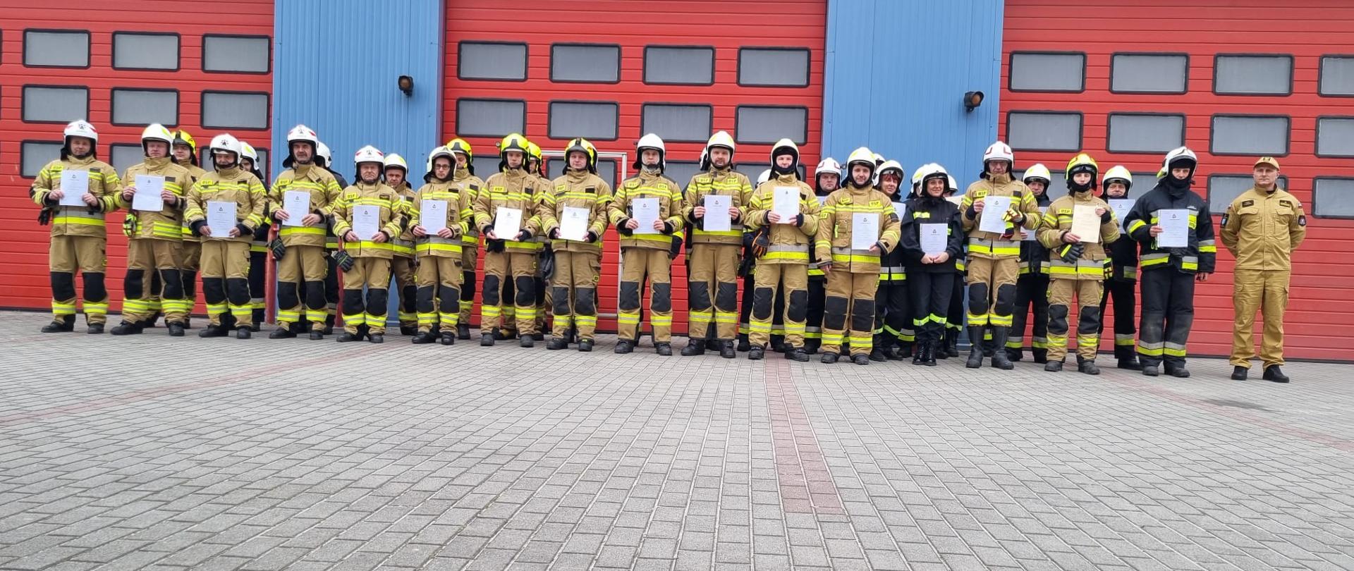 Zdjęcie przedstawia strażaków w umundurowaniu specjalnym na placu komendy powiatowej PSP w Jędrzejowie. Strażacy trzymają w rękach dokumenty potwierdzające ukończenie szkolenia podstawowego.