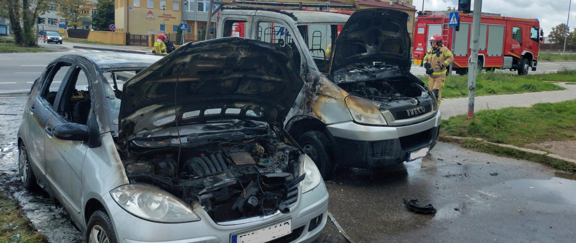 Zdjęcie przedstawia dwa auta, które uległy spaleniu. Za pojazdami widać dwa wozy strażackie.