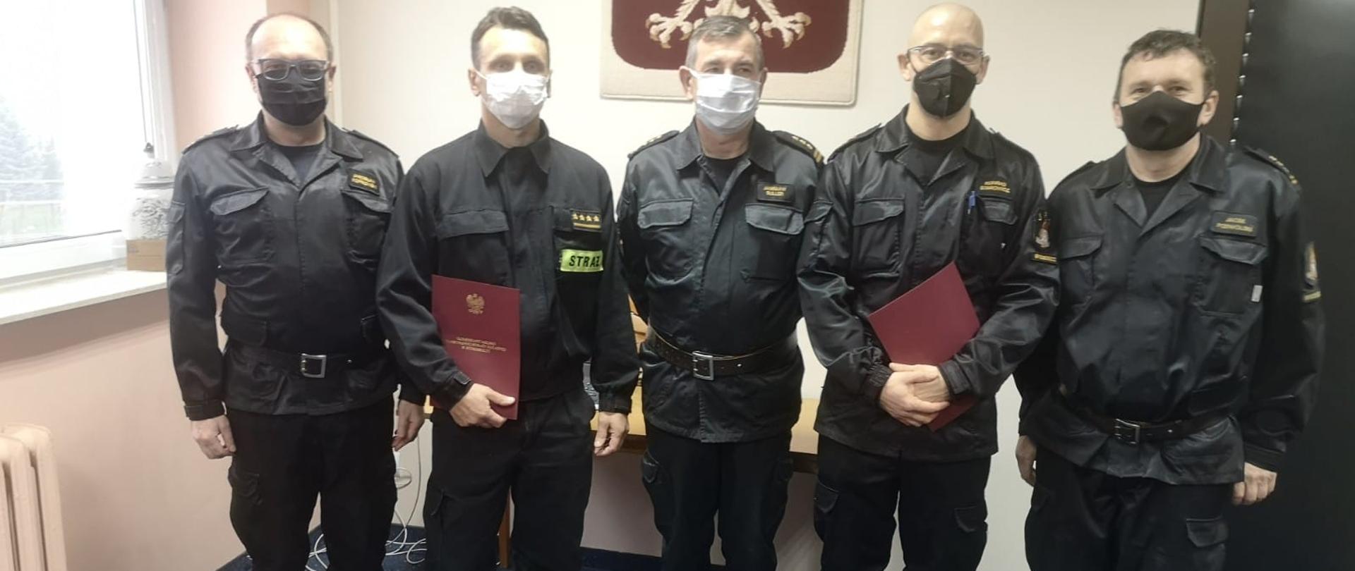 Na zdjęciu st. bryg. Janisław Buller - Komendant Miejski PSP w Bydgoszczy, bryg. Jarosław Jakub Koprowski - Zastępca Komendanta Miejskiego PSP w Bydgoszczy, st. bryg. Jacek Podwolski - Zastępca Komendanta Miejskiego PSP w Bydgoszczy, st. kpt. Konrad Starowicz oraz st. kpt. Krzysztof Bąk. Wszyscy w czarnych ubraniach dowódczo-sztabowych oraz koszarowych, na twarzach maseczki. Wszyscy znajdują się w świetlicy Komendy Miejskiej PSP w Bydgoszczy, w tle Godło Polski na ścianie. 