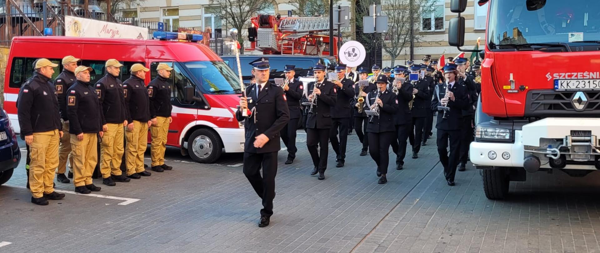 Orkiestra prowadzona jest przez kapelmistrza, w tle znajdują się strażacy oraz samochody strażackie.