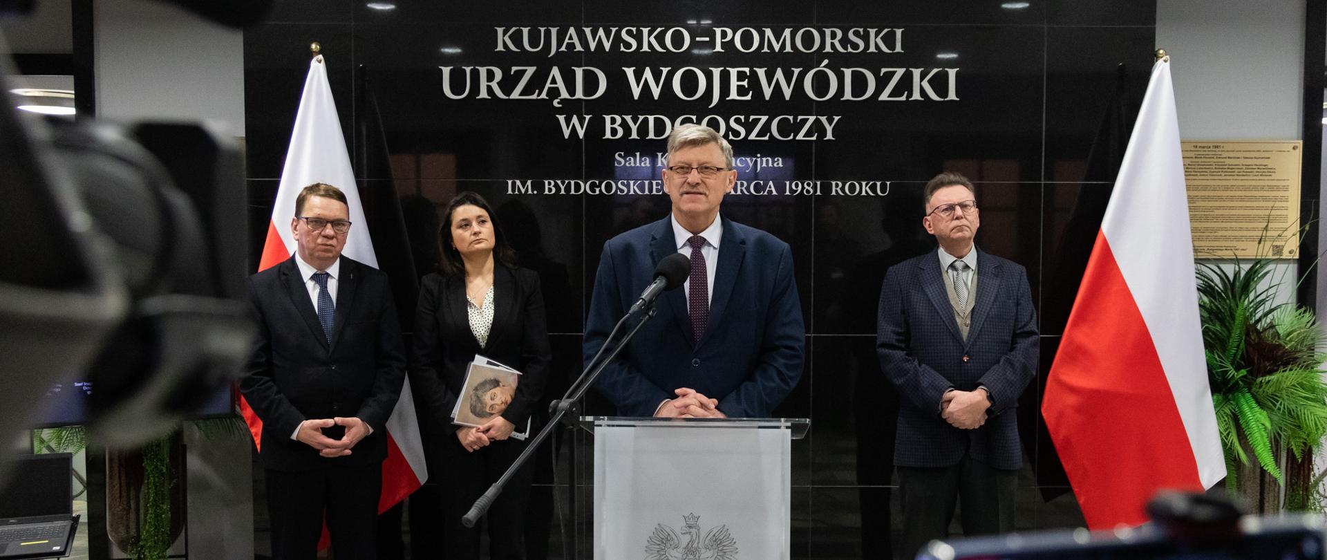 Jeden mężczyzna stojący przy mównicy i wypowiadający się. Za nim kobieta i dwóch mężczyzn