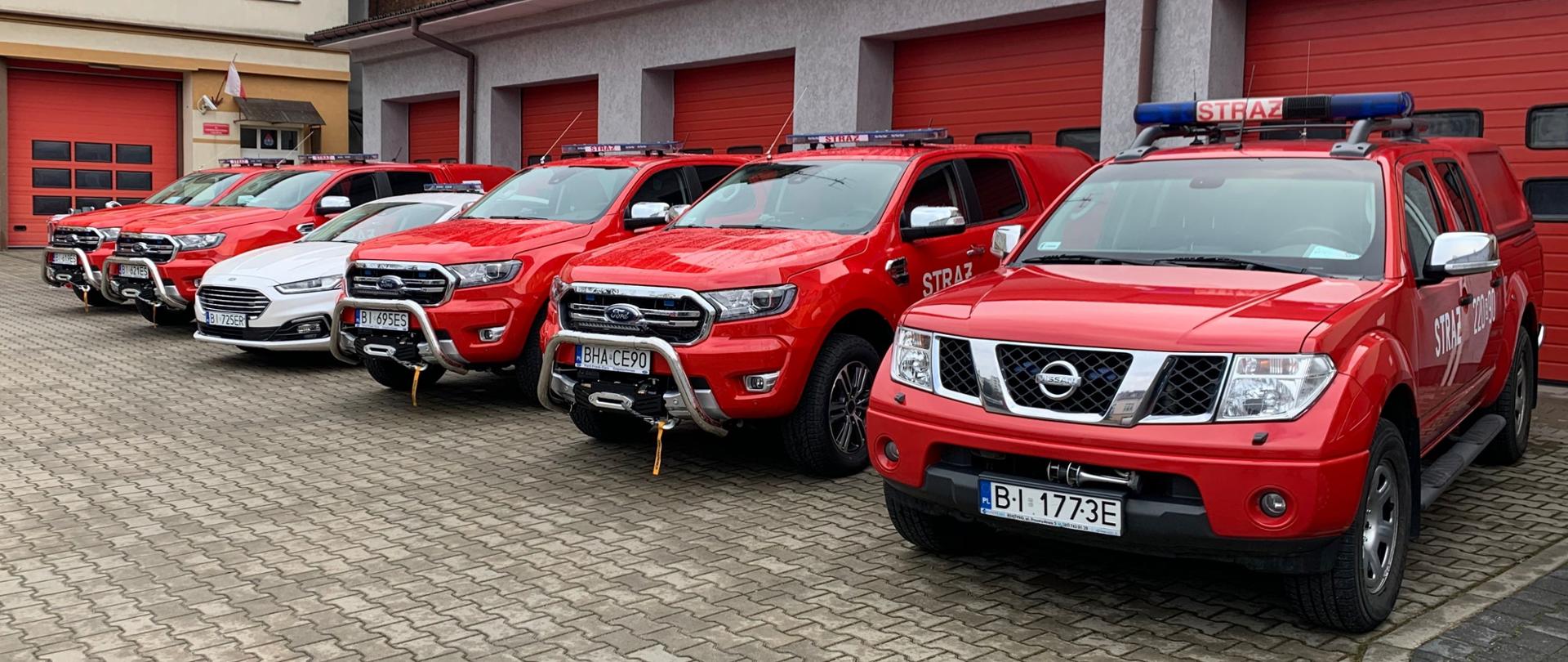 Sześć samochodów pożarniczych ustawionych w szeregu na tle budynku garażowego podczas uroczystości przekazania w/w pojazdów do komend miejskich i powiatowych z terenu województwa podlaskiego.