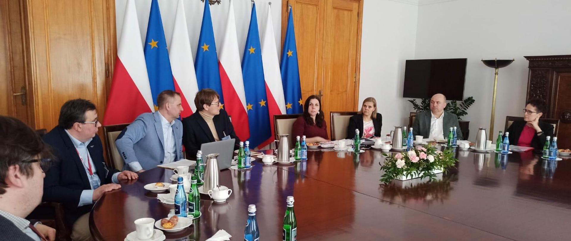 Szefowa służby cywilnej rozmawia z przedstawicielami Fundacji Alumni KSAP przy dużym stole, za nimi znajdują się flagi Polski i Unii Europejskiej.