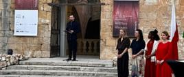 A man dressed in black is speaking on the steps in front of the building. To the right, four women stand in a row: a curator and three artists.