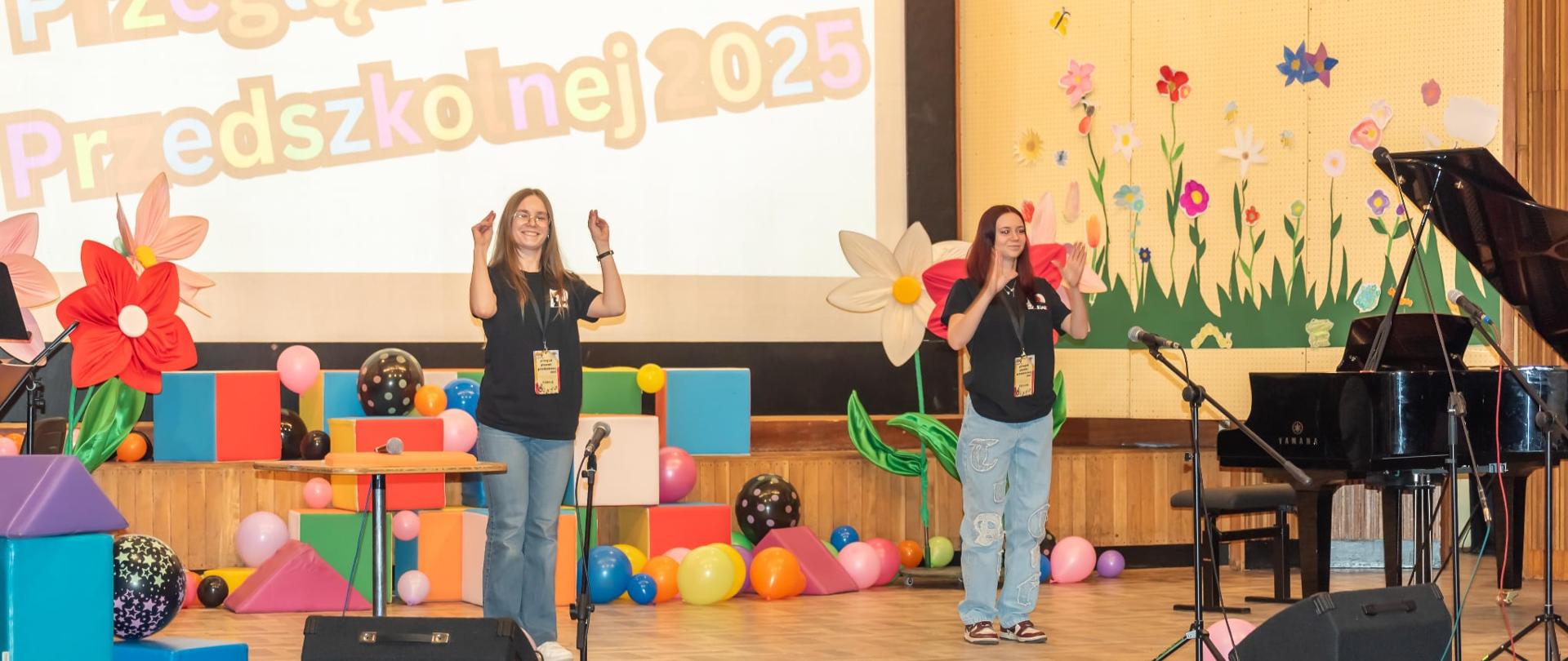 Zdjęcie przedstawia sceną na której stoją dwie słuchaczki Studium. Są tam również kolorowe kostki, balony, papierowe kwiaty. Na ekranie wyświetlany jest napis: Przegląd Piosenki Przedszkolnej