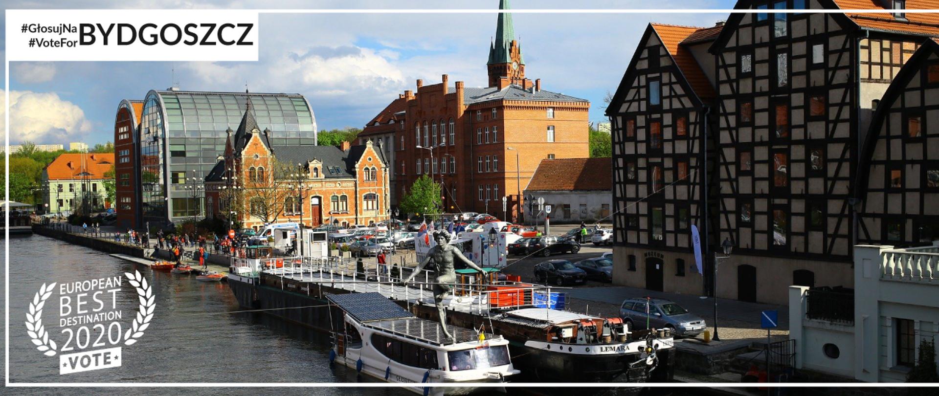 Na zdjęciu panorama Bydgoszczy na Brdą - zabudowania na lądzie, łodzie na wodzie.
Na górze zdjęcia podpis #GłosujNaBydgoszcz, #VoteForBydgoszcz.