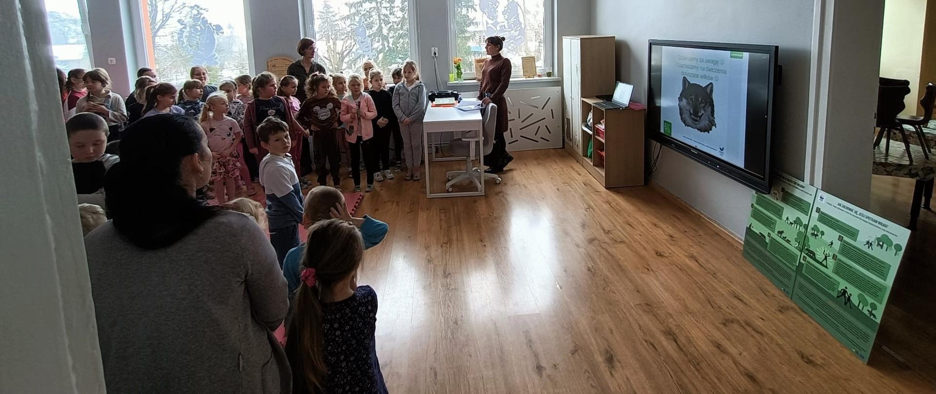 Na zdjęciu grupa uczniów słuchających wykładu na temat wilka. Zostaje wyświetlony wilk na prezentacji. Prelekcję prowadzi pracownik RDOŚ, który stoi przy biurku