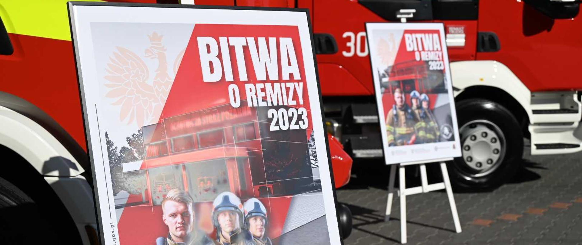 Dwa plakaty promocyjne akcji "Bitwa o Remizy 2023" na tle dwóch pojazdów pożarniczych