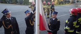 Podniesienie Flagi na maszt z okazji 103 rocznicy odzyskania niepodległości. Poczet flagowy w umundurowaniu galowym. W tle zbiórka strażaków w umundurowaniu bojowym.