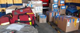 Kilkanaście pudeł kartonowych stojących na sobie leży na garażu, po lewej stronie trzy czerwone torby medyczne, na pierwszym planie węże strażackie w torbach foliowych