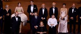 Ministra Edukacji Barbara Nowacka przemawia do mikrofonu ze sceny podczas gali. Za nią stoją uroczyście ubrani goście i gospodarze wydarzenia w garniturach i sukniach wieczorowych.