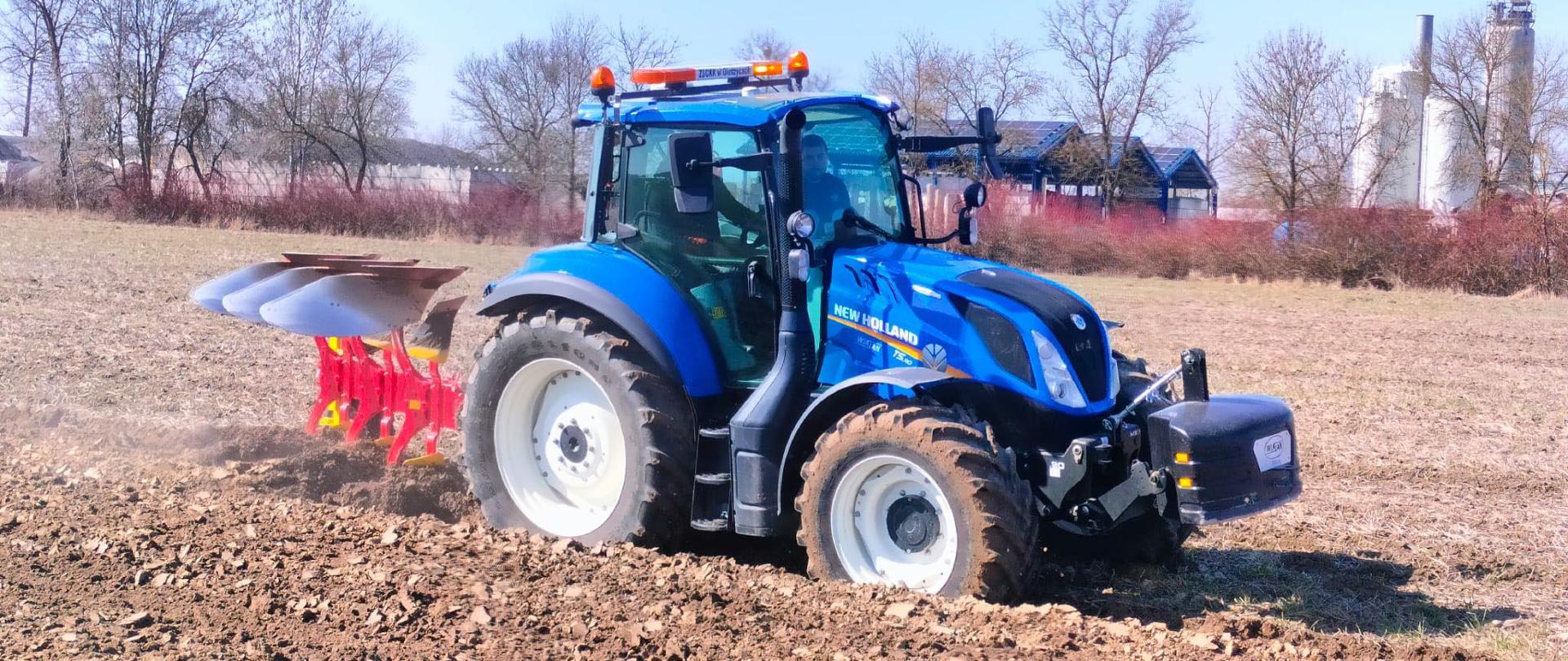 Niebieski ciągnik ze sprzętem rolniczym (pługiem) w trakcie pracy polowej. W tle wiaty i silosy