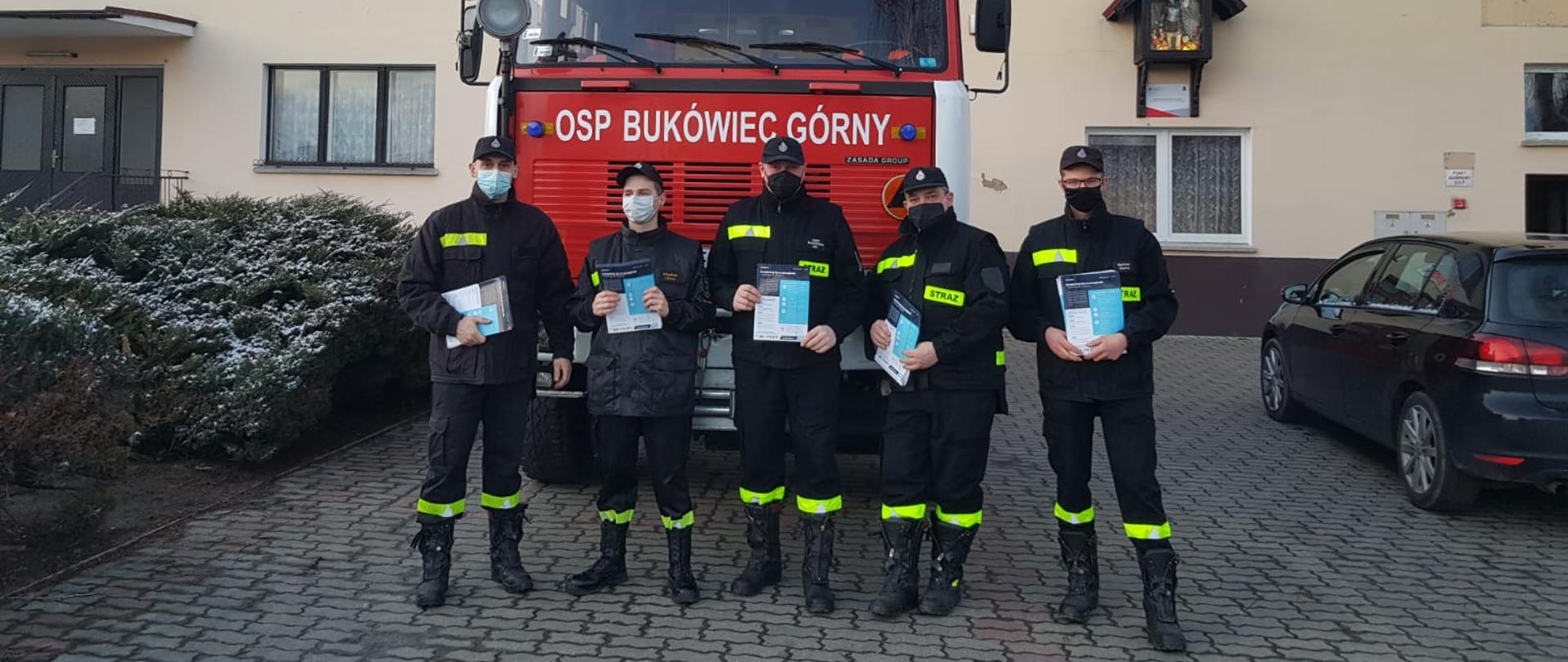 Strażacy stoją na tle samochodu pożarniczego. W rękach trzymają ulotki dot. szczepień.