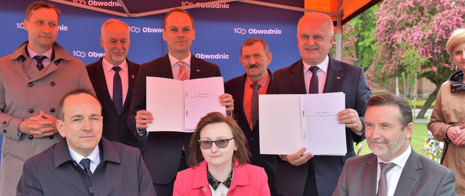 Przy stole siedzą przedstawiciele GDDKiA oraz wykonawcy robót Za nimi stoją uczestnicy uroczystości w tym wojewoda Dajczak, minister Weber, którzy prezentują dokumenty. W tle lokalni samorządowcy 