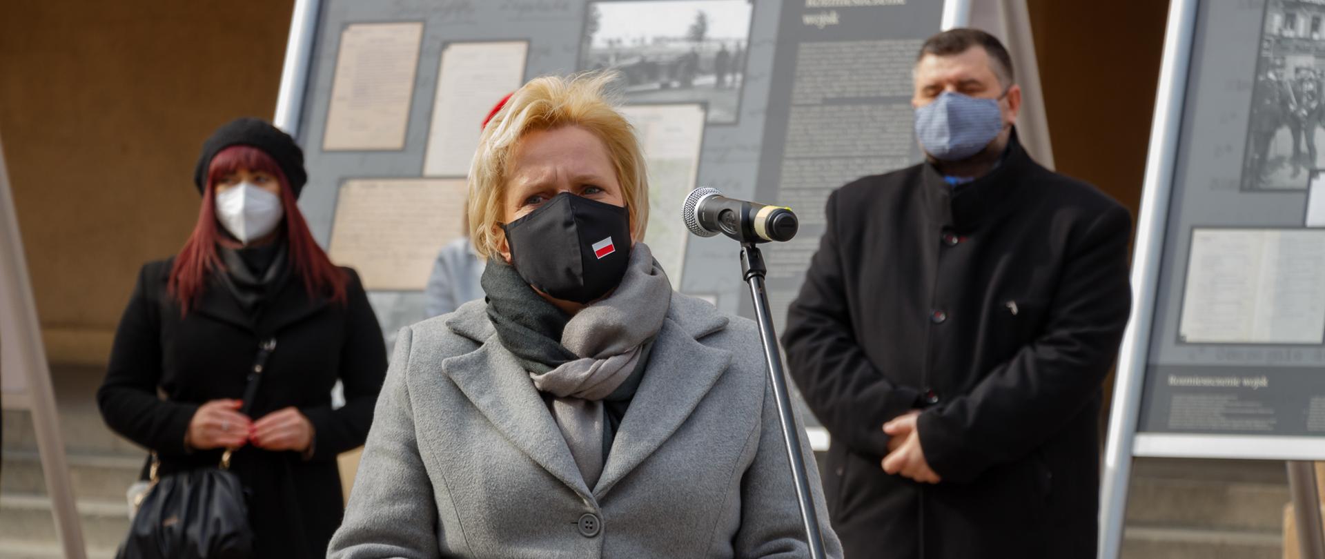 Na zdjęciu przemawiająca przez mikrofon na statywie wicewojewoda opolski Teresa Barańska. W dłoniach trzyma zrulowaną starą gazetę - ulotkę dot. wystawy. Za nią w szeregu stoją trzy osoby. W dalszym planie wystawa na statywach. 