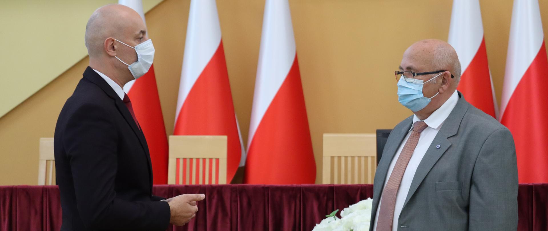 Wojewoda opolski, który znajduje się po lewej stronie kadru wręcza Ireneuszowi Sołkowi Złoty Krzyż Zasługi. W tle stół prezydialny a za nim 5 stojących w rzędzie biało-czerwonych flag RP.