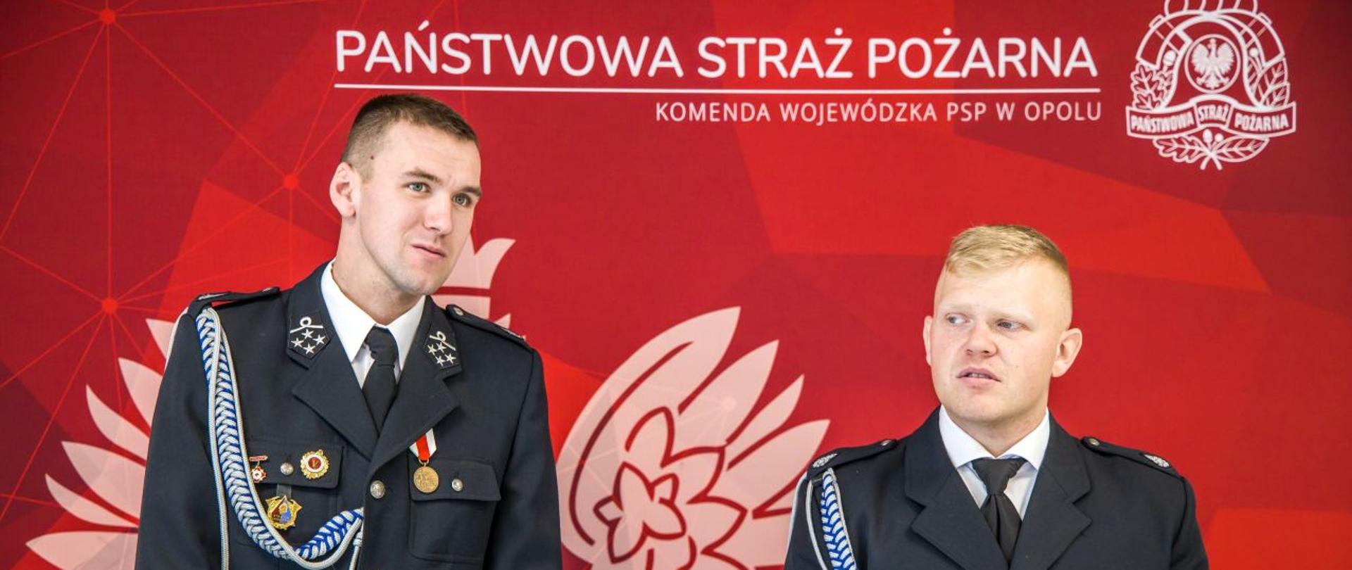 Dh Adam Szczęsny i dh Dawid Jakusz z OSP Osowiec w umundurowaniu wyjściowym stoją na tle ścianki Państwowej Straży Pożarnej koloru czerwonego z logo PSP i Godłem Polski.