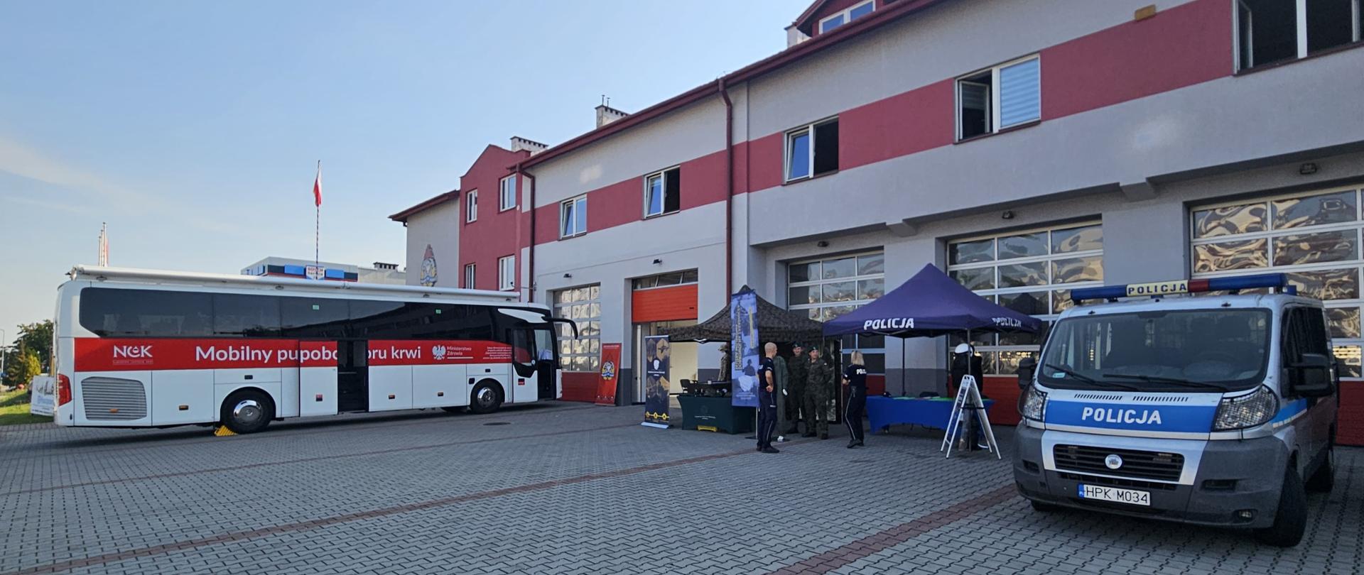 Plac przed budynkiem straży pożarnej, na którym zaparkowany jest biało-czerwony autobus mobilnego punktu krwiodawstwa oraz radiowóz policyjny. Obok rozstawione są namioty policyjne i wojskowe, przy których stoją funkcjonariusze. Na tle budynku widać bramy garażowe i powiewającą flagę państwową.