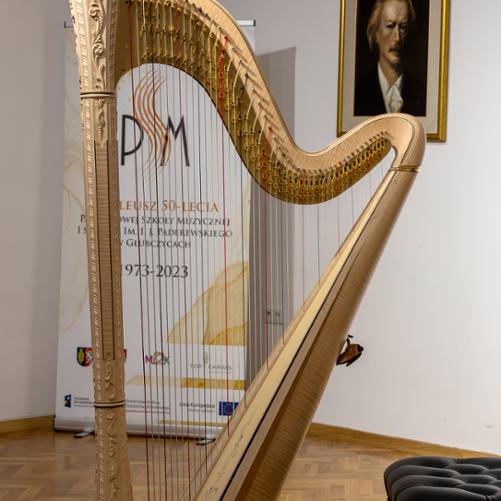 Harfa stojąca na scenie PSM w Głubczycach. W tle widoczny baner szkoły informujący o pięćdziesięcioleciu PSM oraz wiszący na ścianie portret patrona szkoły.