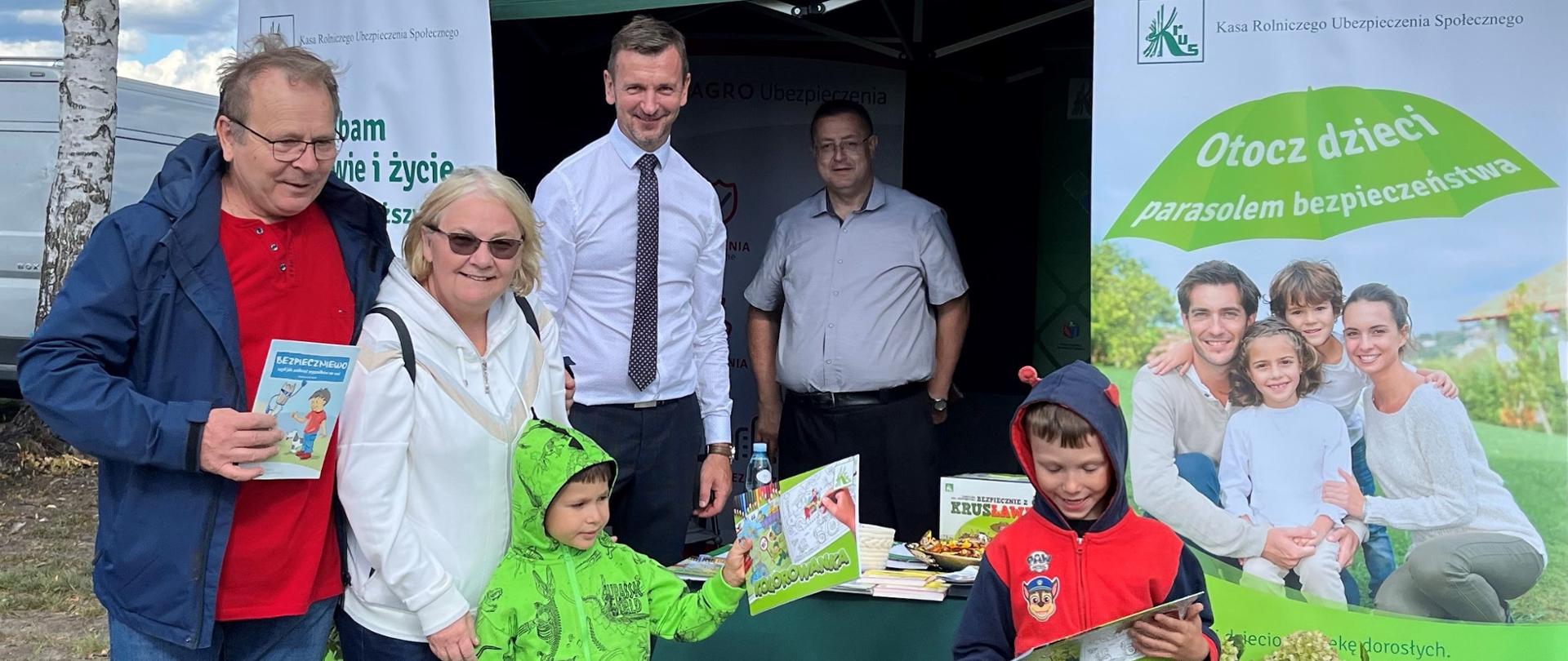 Mężczyzna i kobieta z dwójką dzieci stoją przy stoisku informacyjnym KRUS. Dzieci trzymają w rękach materiały edukacyjne KRUS. Wewnątrz namiotu ze stoiskiem stoją dwaj mężczyźni - pracownicy placówki.
