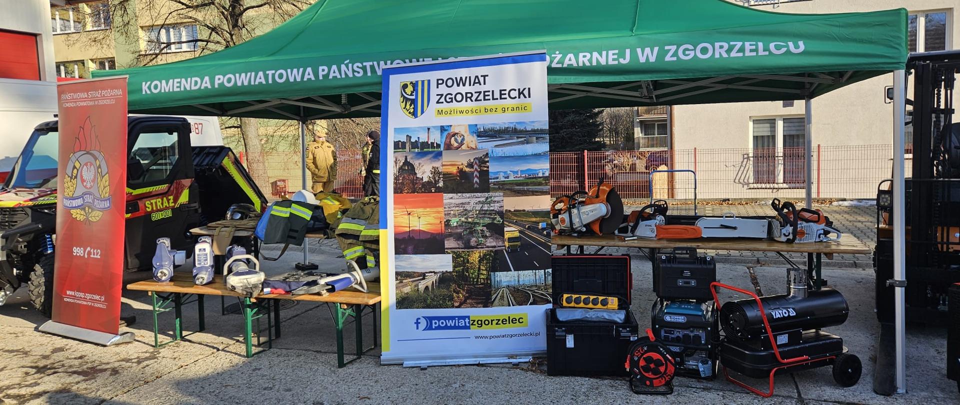 Dzień. Widok na namiot o dachu w kolorze zielonym, bez bocznych ścian. Pod namiotem, na kilku ławkach leży rozłożony różny sprzęt , w tym urządzenia hydrauliczne mundury strażackie pilarki di drewna oraz do stali i betony, nagrzewnice. Z lewej strony namiotu stoi pojazd quad typu UTV, z prawej stoi wózek widłowy. dodatkowo przy namiocie stoją banery informacyjne KP PSP w Zgorzelcu oraz Starostwa Powiatowego w Zgorzelcu.