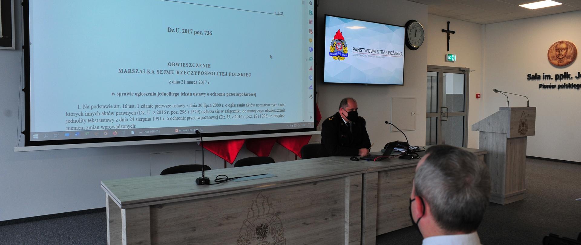 Zastępca pomorskiego komendanta wojewódzkiego PSP omawia projekt ustawy o ochotniczej straży pożarnej. Na ekranie multimedialnym wyświetlany jest projekt ustawy, funkcjonariusz Państwowej Straży Pożarnej słucha wypowiedzi.