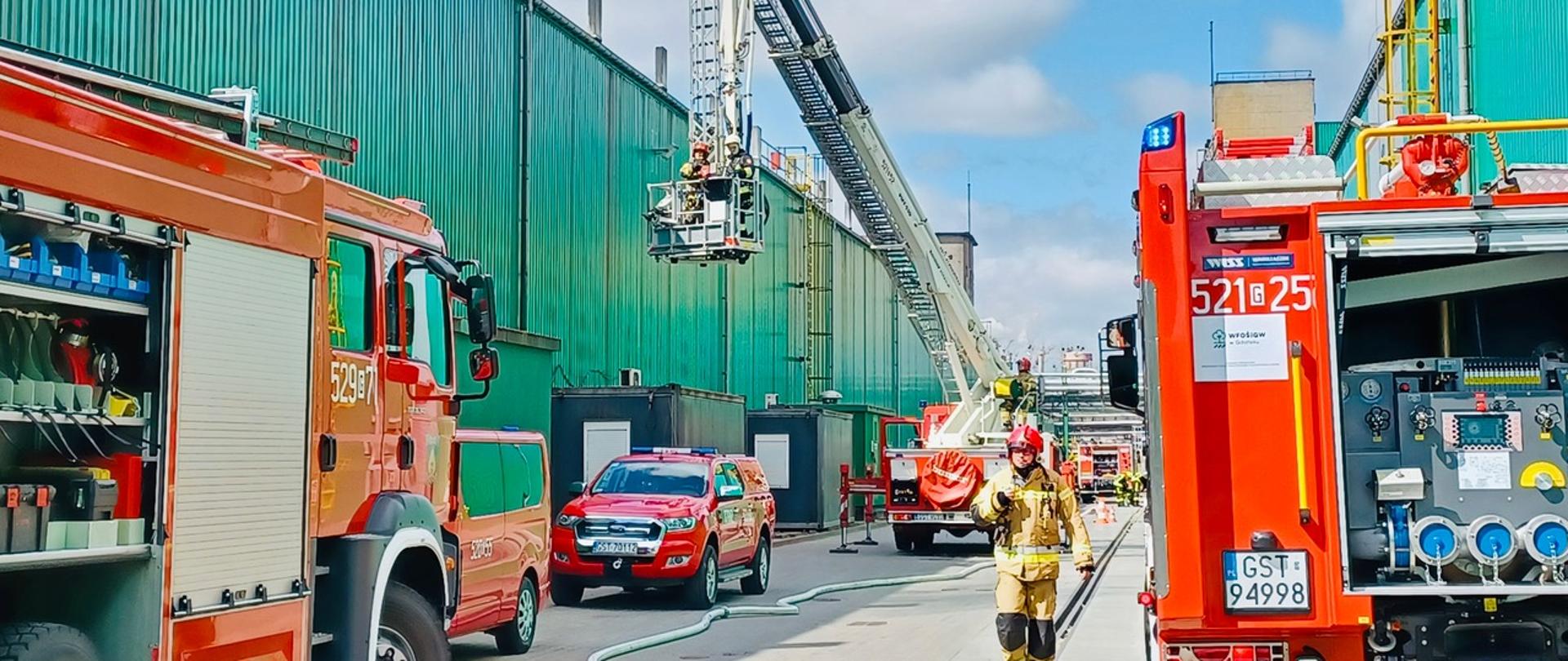 Zdjęcie przedstawia samochody pożarnicze przed zakładem w trakcie ćwiczeń. Na pierwszym planie strażak. W tle zakład produkcyjny