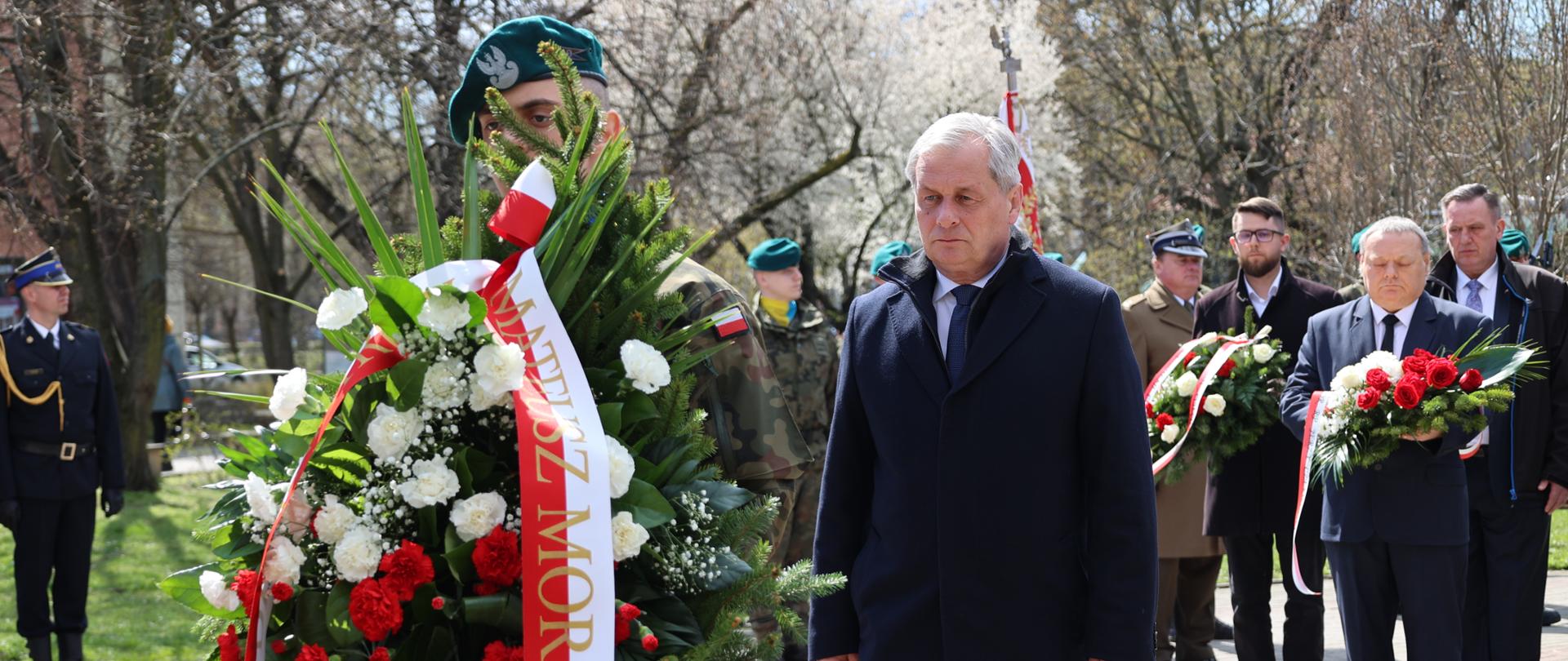 Mężczyzna ubrany na czarno zmierza za żołnierzem z wieńcem premiera RP