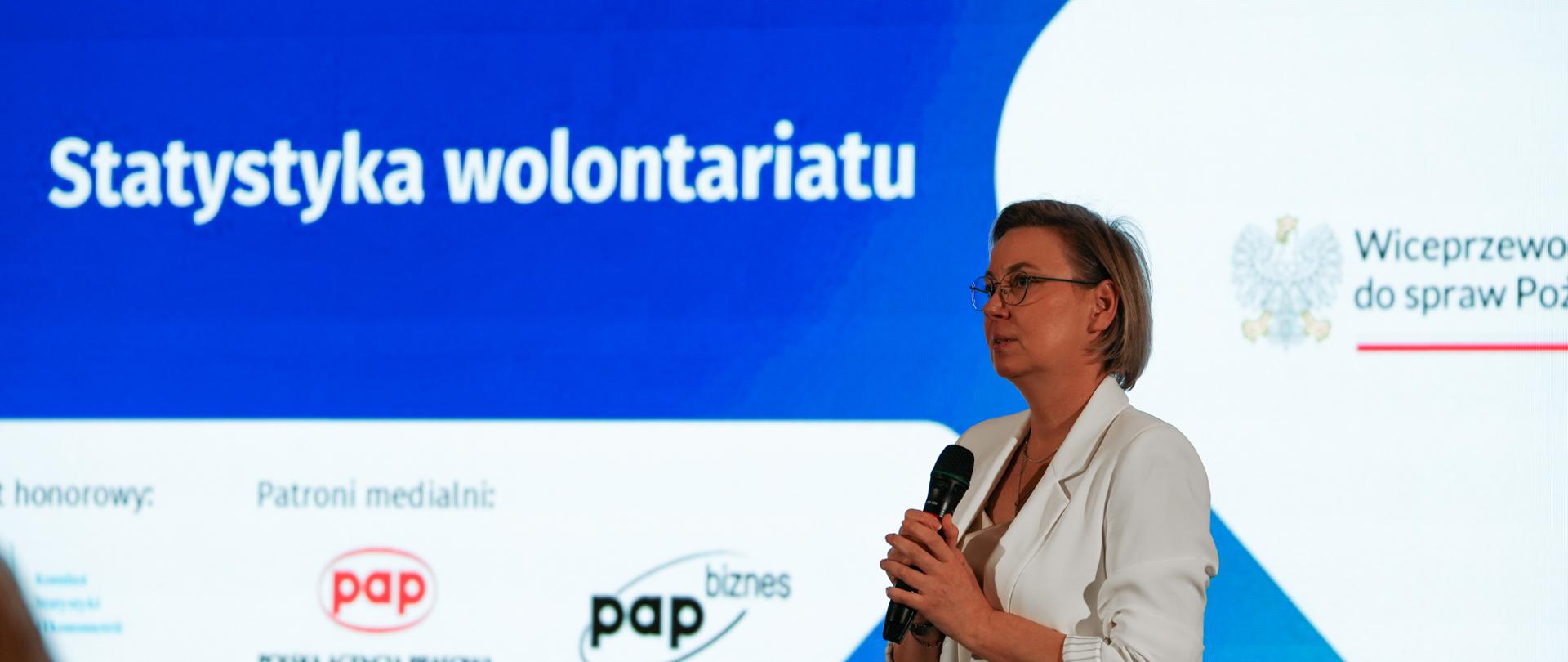 Zdjęcie sytuacyjne. Pani Adriana Porowska, Sekretarz Stanu w Kancelarii Prezesa Rady Ministrów, Wiceprzewodnicząca Komitetu do spraw Pożytku Publicznego przemawia do uczestników konferencji, trzymając mikrofon w dłoniach. Za Panią Wiceprzewodniczącą widoczny jest ekran, na którym wyświetlony jest tytuł: Statystyka wolontariatu. Na ekranie widoczne są loga patronów medialnych pap i pap biznes oraz fragment loga Wiceprzewodniczącej Komitetu do spraw Pożytku Publicznego.