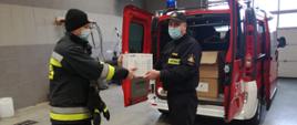 strażak przekazuje paczkę z środkami do walki z SARS, w tle samochód czerwony pożarniczy, zdarzenie ma miejsce w garażu KM PSP w Płocku