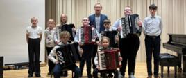 Grupa dzieci i nastolatków pozuje na scenie sali koncertowej z akordeonami. W centralnej części stoi dorosły opiekun. Część uczniów trzyma akordeony w rękach, a dwoje siedzi na krzesłach z instrumentami na kolanach. W tle znajdują się jasne, pionowe panele akustyczne oraz fortepian po prawej stronie. Wszyscy są ubrani odświętnie i patrzą w stronę aparatu.