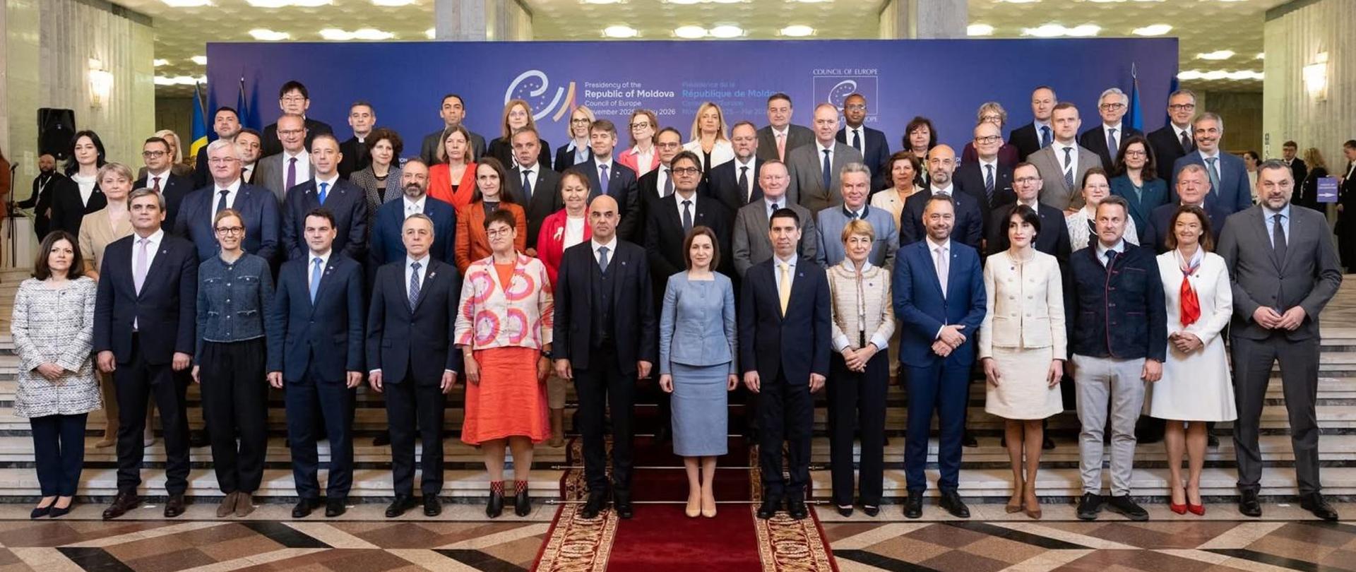 Na zdjęciu jest grupa ok. 20 osób. Stoją i patrzą w kierunku obiektywu. Za nimi jest niebieska ścianka z napisem "Republic of Moldova".