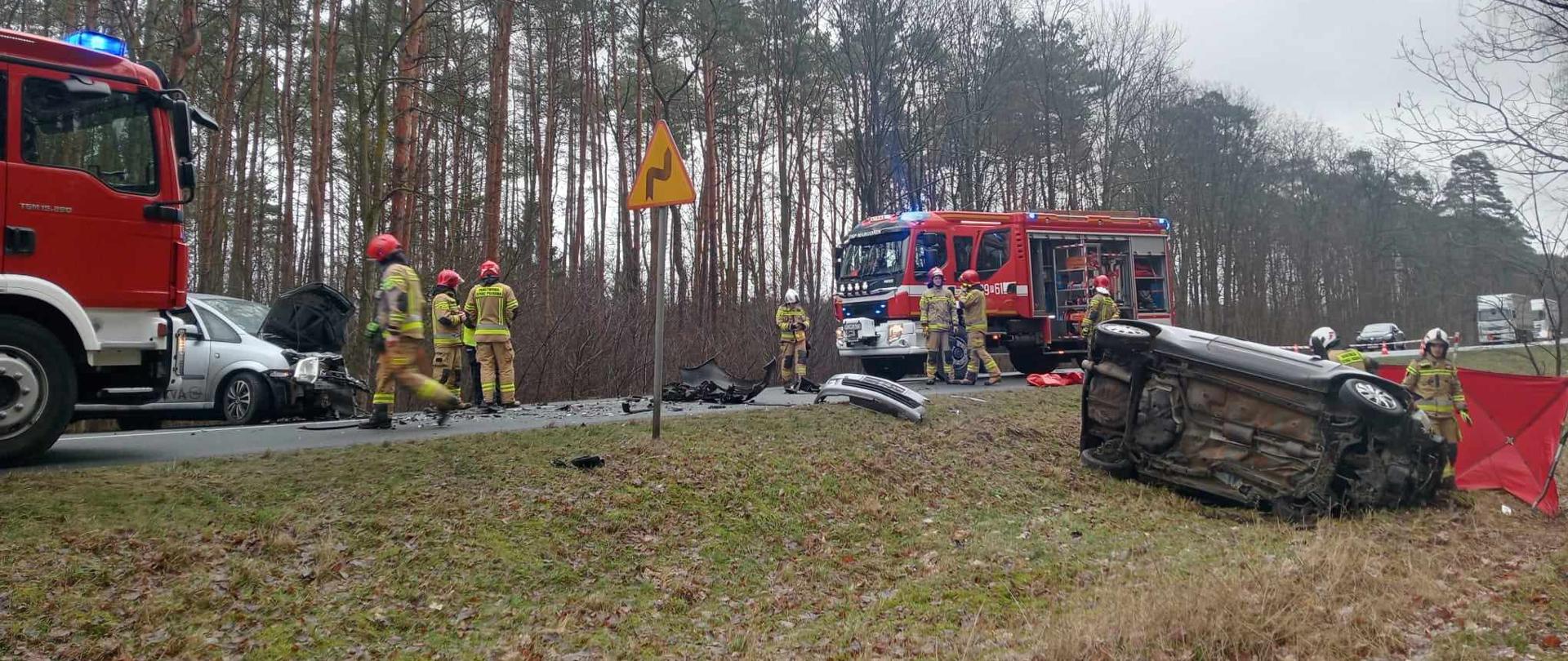 Zdjęcie przedstawia strażaków i policjantów działających przy wypadku drogowym.
W tle pojazdy uszkodzone, samochody strażackie i las.
