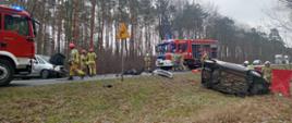 Zdjęcie przedstawia strażaków i policjantów działających przy wypadku drogowym.
W tle pojazdy uszkodzone, samochody strażackie i las.
