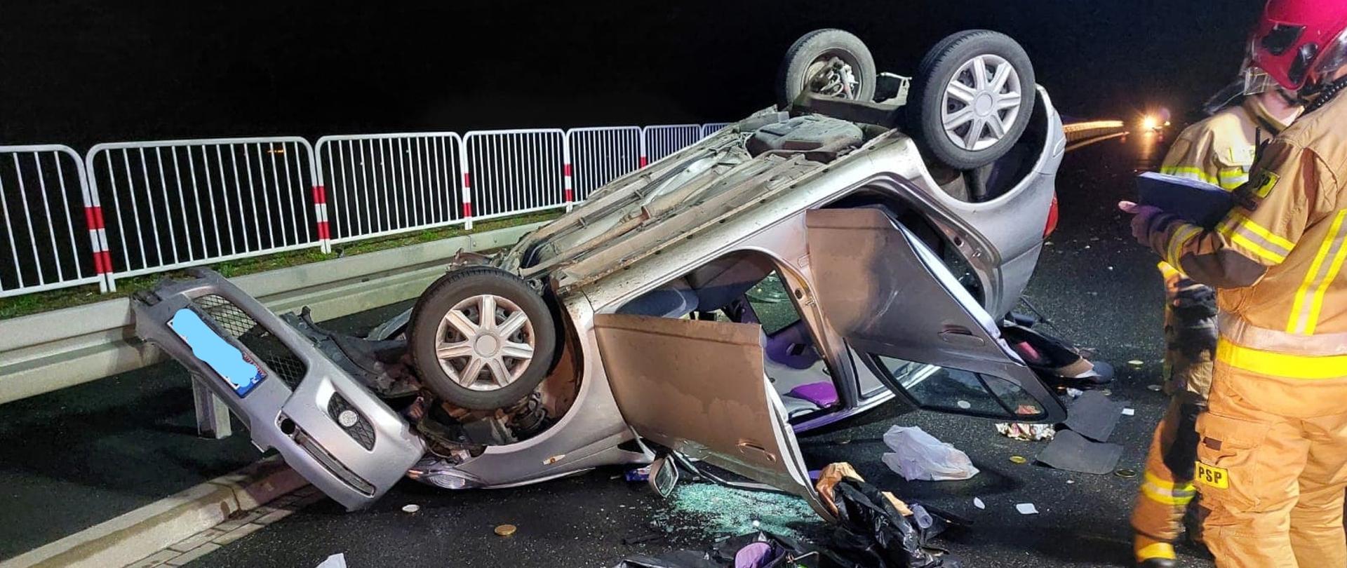 Zdjęcie przedstawia rozbity samochód osobowy leżący na dachu. Na jedni rozrzucone części i przedmioty. Po prawej stronie stoi dwóch strażaków