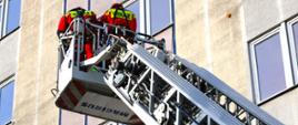 Dwaj strażacy w koszu drabiny, w tle wysoki budynek