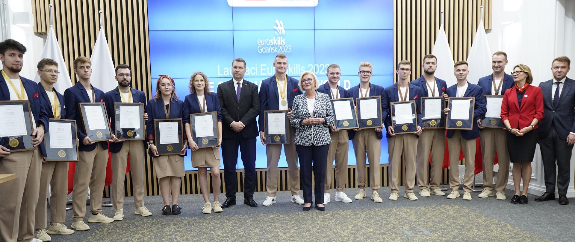 W dużej sali pod ścianą stoi duża grupa trzymających dyplomy młodych ludzi w granatowych marynarkach, przed nimi minister Czarnek i wiceminister Machałek.