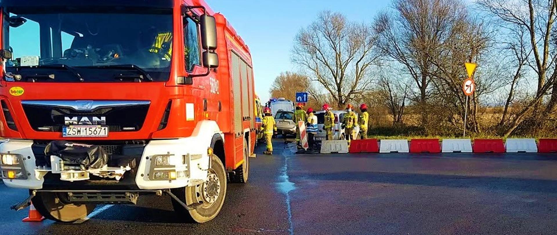 Skrzyżowanie ulic. Na pierwszym planie po lewej przodem stoi średni samochód pożarniczy, na dalszym planie stoją dwa rozbite srebrne samochody osobowe oraz karetka ZRM w kolorze żółtym. Strażacy w umundurowaniu specjalnym zabezpieczają miejsce zdarzenia.
