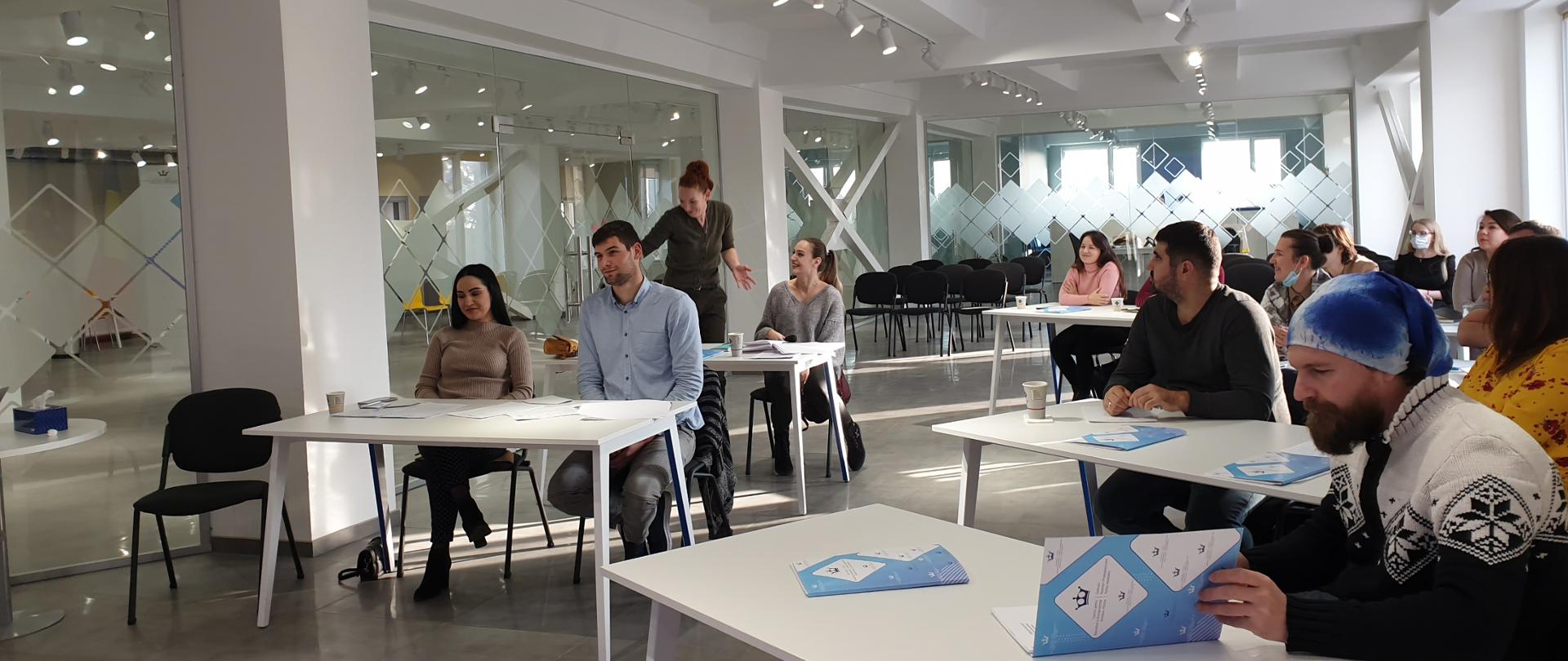 Grupa młodych ludzi siedzi w ławkach w jasnej, przestrzennej, oszklonej sali.