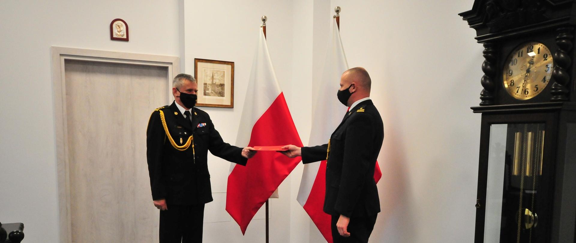 Pomorski Komendant Wojewódzki Państwowej Straży Pożarnej starszy brygadier Piotr Socha wręcza w Komendzie Wojewódzkiej Państwowej Straży Pożarnej w Gdańsku awans na stopień młodszego brygadiera Piotrowi Krzemińskiemu Komendantowi Powiatowemu Państwowej Straży Pożarnej w Lęborku. W tle widoczne flagi narodowe. Z powodu pandemii koronawirusa wszyscy mają czarne maseczki ochronne na twarzy.