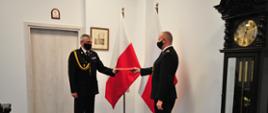 Pomorski Komendant Wojewódzki Państwowej Straży Pożarnej starszy brygadier Piotr Socha wręcza w Komendzie Wojewódzkiej Państwowej Straży Pożarnej w Gdańsku awans na stopień młodszego brygadiera Piotrowi Krzemińskiemu Komendantowi Powiatowemu Państwowej Straży Pożarnej w Lęborku. W tle widoczne flagi narodowe. Z powodu pandemii koronawirusa wszyscy mają czarne maseczki ochronne na twarzy.