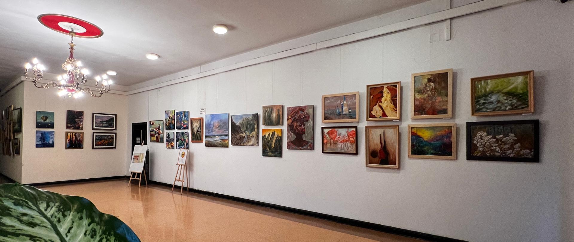 Panorama części wystawy. Obrazy wiszą na białej ścianie. Na suficie żyrandol i lampy punktowe.